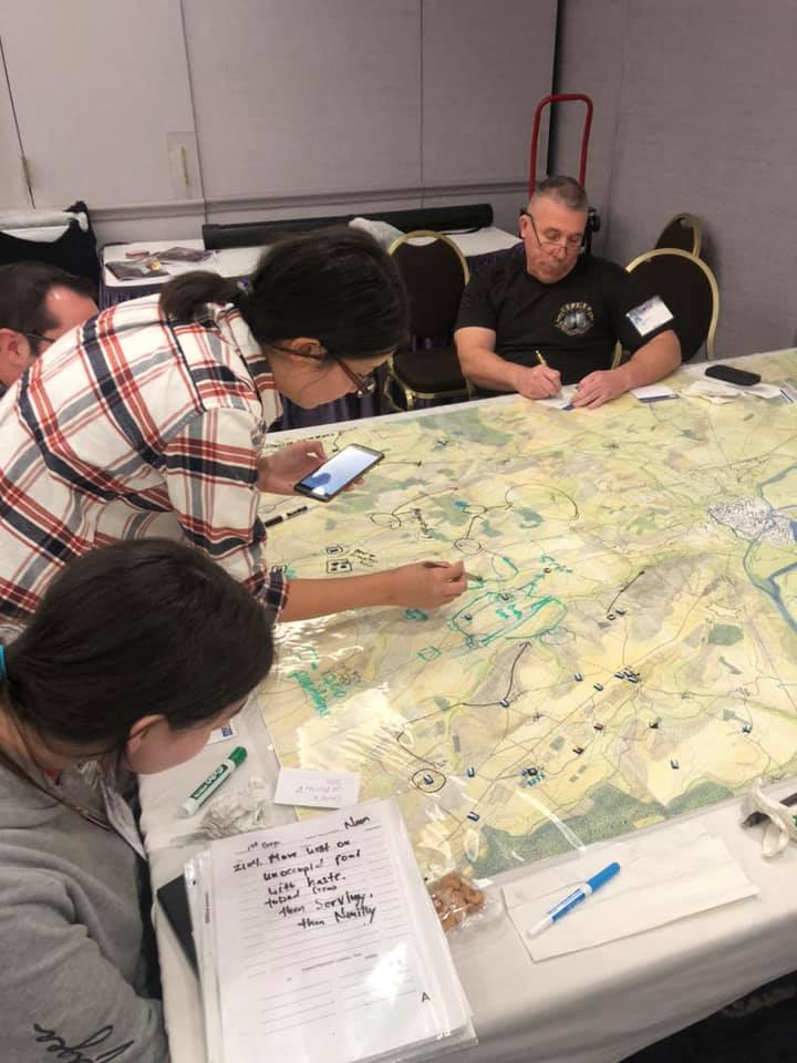 Women and men sitting and standing around atable with a huge map, writing orders and drawing troop movements onto a plastic layer on top of the map.